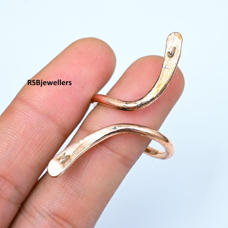 May include: A rose gold-coloured, twisted, open-ended ring. The ring is made of metal and has a smooth, polished finish. The ring is marked with the letter "M" on one side and the letter "J" on the other side.