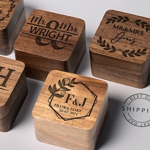 May include: Five wooden ring boxes with engraved personalized messages. The boxes are made of dark wood and have a square shape. The engravings include names, dates, and initials.