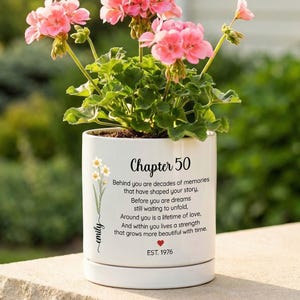 May include: A white ceramic planter containing pink geranium flowers. The planter features the text 'Chapter 50', a poem about memories and dreams, the name 'emily' written vertically with a flower illustration, a small red heart, and 'EST. 1976'.