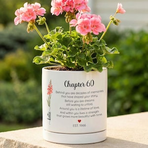 May include: A white ceramic planter with a saucer containing a pink geranium plant. The planter features the text "Chapter 60" followed by a poem about memories and dreams, a small red heart, the name "Katie", and the text "EST. 1966".