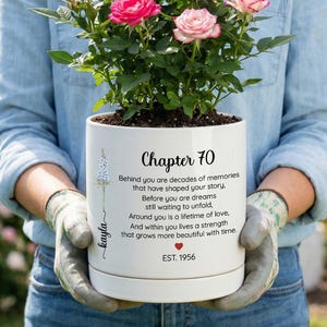 May include: A white ceramic planter containing a miniature rose bush with pink flowers. The planter features black printed text that reads: Chapter 70, Behind you are decades of memories that have shaped your story, Before you are dreams still waiting to unfold, Around you is a lifetime of love, And within you lives a strength that grows more beautiful with time, EST. 1956, and the name Kayla.