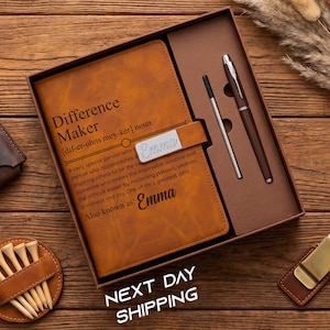 May include: A brown leather journal set with a pen, a small pen, and a money clip. The journal has the text "Difference Maker" and the name "Emma". The set is in a gift box, with a golf tee holder and a small leather wallet.