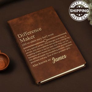 May include: A brown leather-bound journal featuring the gold-embossed text Difference Maker, a phonetic pronunciation guide, a definition of the term, and the name James. The cover has a smooth texture and a rectangular shape with rounded corners.