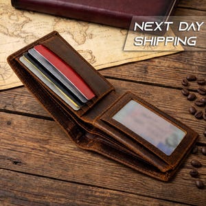 May include: A brown leather bifold wallet featuring multiple card slots containing red, black, and gray cards, along with a clear plastic ID window. The wallet is open, displaying its interior compartments and stitching details on the leather surface.