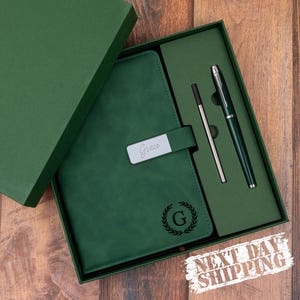 May include: A green journal and pen set in a gift box. The journal has a silver nameplate that reads "Grace" and a "G" with a laurel wreath. The set includes a pen and a stylus. The box is green and has a "Next Day Shipping" sticker.