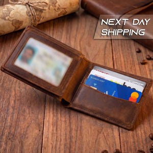 May include: A brown leather bi-fold wallet featuring a clear plastic ID window on the left side and multiple card slots on the right side. The wallet is open, displaying a blue debit card and other cards tucked into the interior pockets.