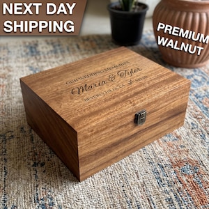 May include: A rectangular walnut wood keepsake box with a hinged lid and a silver metal latch. The lid is engraved with the text "OUR WEDDING MEMORIES", "Maria & Tyler", "5.12.20", and "BEVERLY HILLS, CA" in a script and serif font.