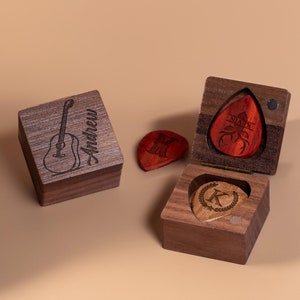 May include: Two wooden boxes with engraved guitar picks. One box is closed and has the name "Andrew" engraved on it. The other box is open and has a guitar pick with the letter "K" engraved on it. The box also has a guitar pick with a deer and the name "Kevin" engraved on it.