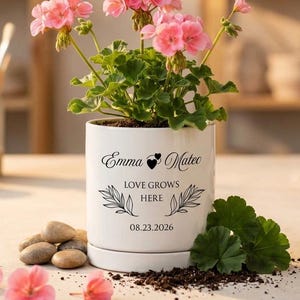 May include: A white ceramic planter containing a pink geranium plant. The planter features black text reading Emma and Mateo, Love Grows Here, and the date 08.23.2026, with a small heart icon and leaf illustrations printed on the front surface.