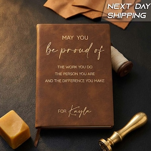 May include: A brown leather-textured journal with gold embossed text that reads: MAY YOU be proud of THE WORK YOU DO THE PERSON YOU ARE AND THE DIFFERENCE YOU MAKE FOR Kayla. A gold wax seal stamp and a block of yellow wax are placed nearby.