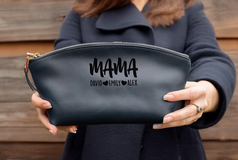 May include: Black leather makeup bag with a zipper closure. The bag has the word "MAMA" written in a stylized font with the names "David", "Emily", and "Alex" written below it, each name separated by a heart.