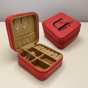 Puede incluir: Caja de joyer&iacute;a de imitaci&oacute;n de cuero rojo con cierre de cremallera. La caja est&aacute; abierta y tiene compartimentos para anillos, pendientes y collares. La caja tiene un dise&ntilde;o floral con la letra "U" en la parte superior.