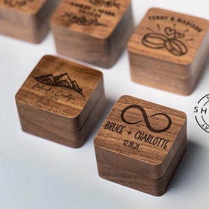 May include: Five wooden ring boxes with engraved designs. The boxes are square and have a dark wood finish. The designs include mountains, an infinity symbol, and two rings.