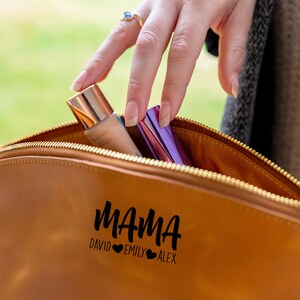 May include: A brown leather makeup bag with a zipper closure. The bag has the word "MAMA" written on it in black cursive lettering, with the names "DAVID", "EMILY", and "ALEX" written below in smaller black lettering. There are two hearts between each name.