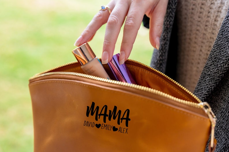 May include: A brown leather makeup bag with a zipper closure. The bag has the word "MAMA" written on it in black cursive lettering, with the names "DAVID", "EMILY", and "ALEX" written below in smaller black lettering. There are two hearts between each name.