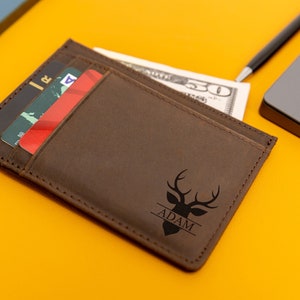 May include: A brown leather wallet with a deer head design and the name "ADAM" engraved on it. The wallet has a slot for cash and multiple card slots.