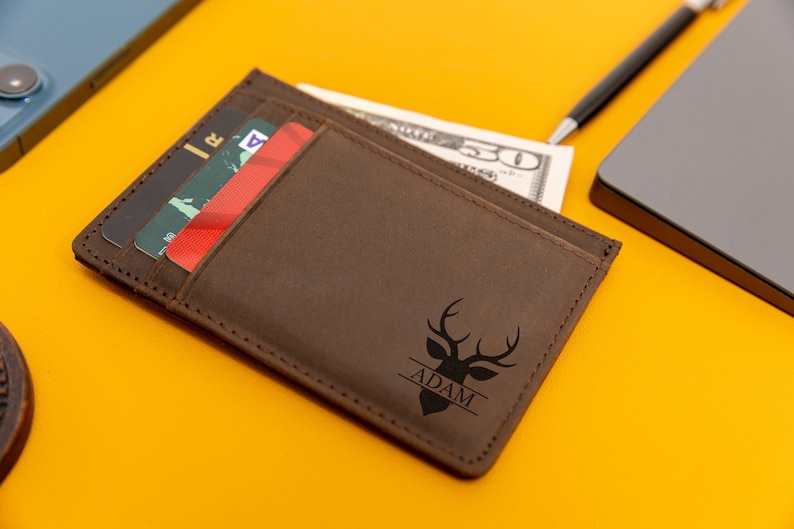 May include: A brown leather wallet with a deer head design and the name "ADAM" engraved on it. The wallet has a slot for cash and multiple card slots.