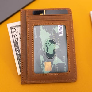 May include: A brown leather wallet with a clear window for displaying a credit card. The card has the VISA logo and the text "10/23" printed on it. The wallet also has a slot for cash or other cards.