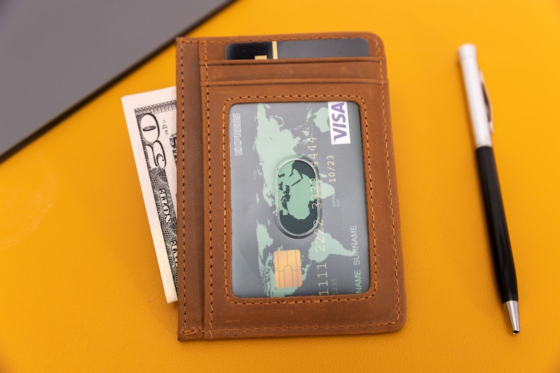 May include: A brown leather wallet with a clear window for displaying a credit card. The card has the VISA logo and the text "10/23" printed on it. The wallet also has a slot for cash or other cards.