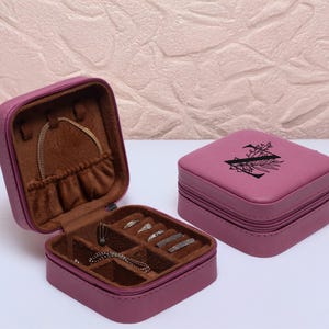 Puede incluir: Una peque&ntilde;a caja de joyer&iacute;a rosa de dos niveles con cierre de cremallera. El nivel superior tiene un dise&ntilde;o floral con la letra "N" en el centro. El nivel inferior tiene compartimentos para anillos, pendientes y collares.