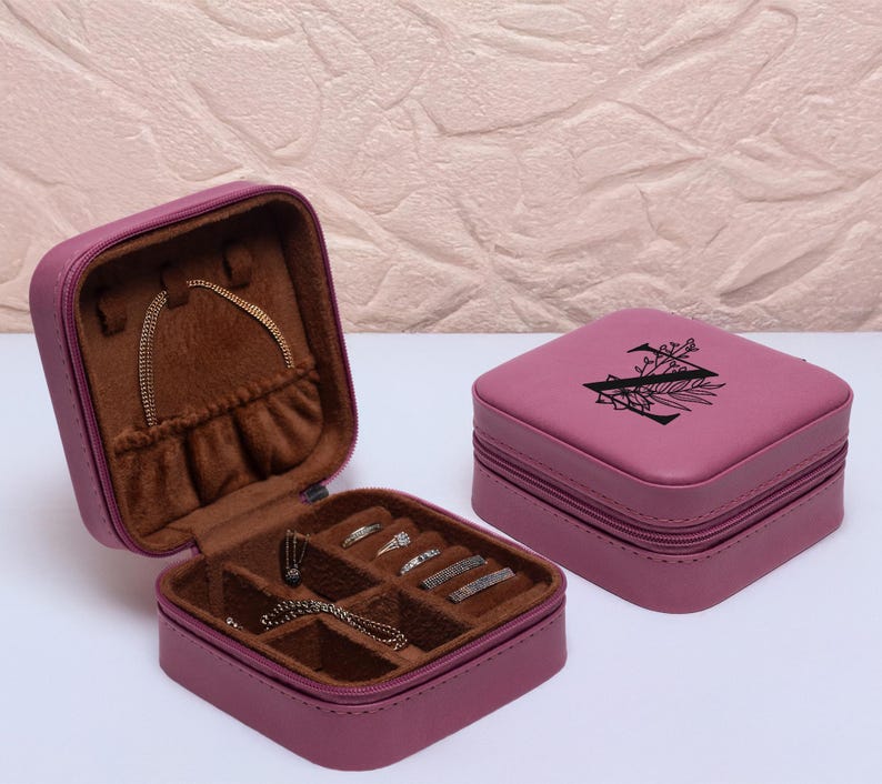 Puede incluir: Una peque&ntilde;a caja de joyer&iacute;a rosa de dos niveles con cierre de cremallera. El nivel superior tiene un dise&ntilde;o floral con la letra "N" en el centro. El nivel inferior tiene compartimentos para anillos, pendientes y collares.