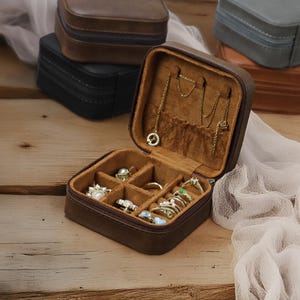 Puede incluir: Una caja de joyer&iacute;a de cuero marr&oacute;n con cierre de cremallera. La caja est&aacute; abierta y tiene varios compartimentos para guardar anillos, pendientes y collares. La caja est&aacute; sobre una superficie de madera.