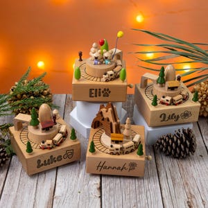 Może przedstawiać: Cztery drewniane pozytywki z motywami pociąg&oacute;w i tor&oacute;w. Każde pudełko ma inną nazwę: Eli, Lily, Luke i Hannah. Pudełka mają miniaturowe drzewa, g&oacute;ry i figurki niedźwiedzi. Pudełko Eli ma odcisk łapy, a pudełko Lily ma kwiat.