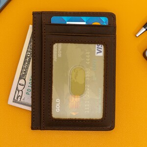 May include: A brown leather wallet with a card slot and a clear window for displaying a credit card. The wallet has the word "GOLD" printed on it. The wallet is on a yellow surface with a dollar bill and a pen.