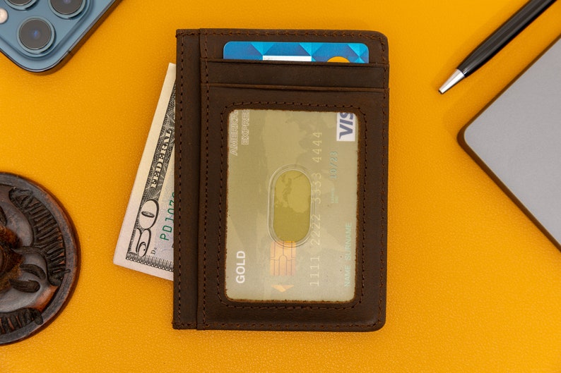 May include: A brown leather wallet with a card slot and a clear window for displaying a credit card. The wallet has the word "GOLD" printed on it. The wallet is on a yellow surface with a dollar bill and a pen.