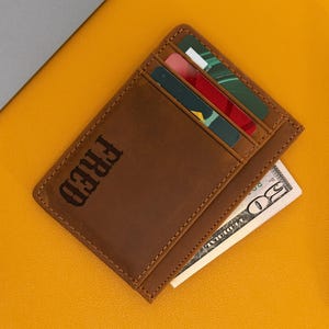 May include: A brown leather cardholder with the name "FRED" embossed on the front. The cardholder contains several credit cards and a fifty-dollar bill. A black pen rests beside the cardholder on a yellow surface.