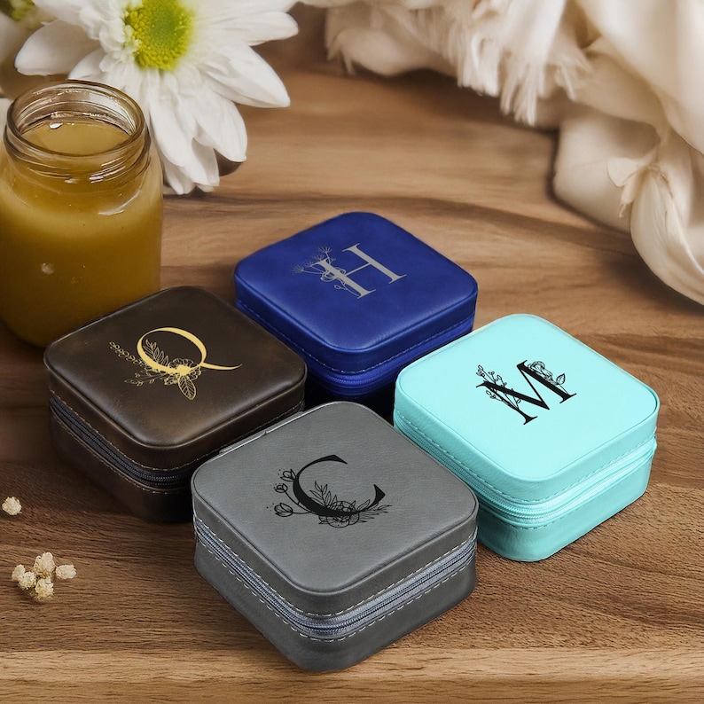 Puede incluir: Cuatro peque&ntilde;as cajas cuadradas de joyer&iacute;a con diferentes exteriores de tela. Cada caja tiene una letra diferente, C, H, M y Q, con un dise&ntilde;o floral en la parte delantera. Las cajas est&aacute;n sobre una superficie de madera.