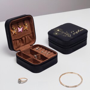 Puede incluir: Caja de joyer&iacute;a negra con un dise&ntilde;o floral y el nombre "Sophia" en la tapa. La caja est&aacute; abierta para revelar un interior marr&oacute;n con compartimentos para anillos y aretes.