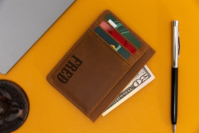 May include: A brown leather wallet with the name "FRED" embossed on the front. The wallet has multiple card slots and a cash pocket. It is lying on a yellow surface with a pen and a $20 bill.