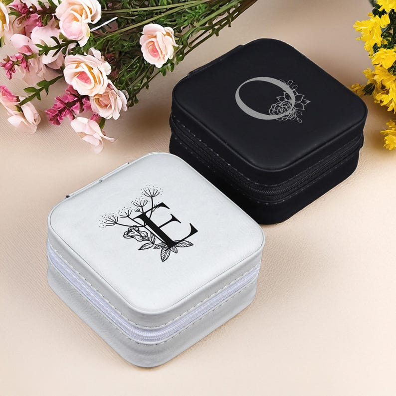 Puede incluir: Dos peque&ntilde;as cajas cuadradas de joyer&iacute;a con cierre de cremallera. La caja de la izquierda es blanca con un dise&ntilde;o floral negro y la letra "E". La caja de la derecha es negra con un dise&ntilde;o floral plateado y la letra "O".