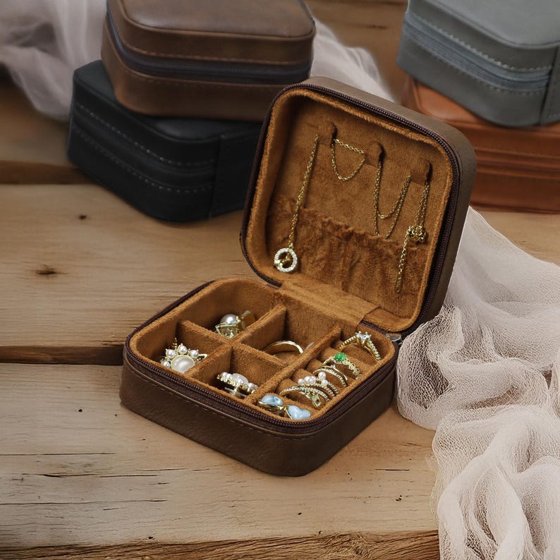 Puede incluir: Una caja de joyer&iacute;a de imitaci&oacute;n de cuero marr&oacute;n con cierre de cremallera. La caja est&aacute; abierta y tiene varios compartimentos para guardar anillos, pendientes y collares. La caja est&aacute; sobre una superficie de madera.
