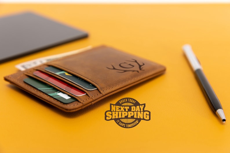 May include: A brown leather wallet with a card slot and a monogrammed initial "G" with antlers. The wallet is on a yellow surface with a pen and a phone.