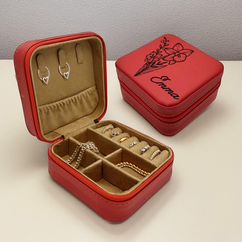 Puede incluir: Caja de joyer&iacute;a de imitaci&oacute;n de cuero rojo con cierre de cremallera. La caja est&aacute; abierta y tiene varios compartimentos para guardar anillos, pendientes y collares. La caja tiene un dise&ntilde;o floral y el nombre "Emma" grabado en la parte superior.