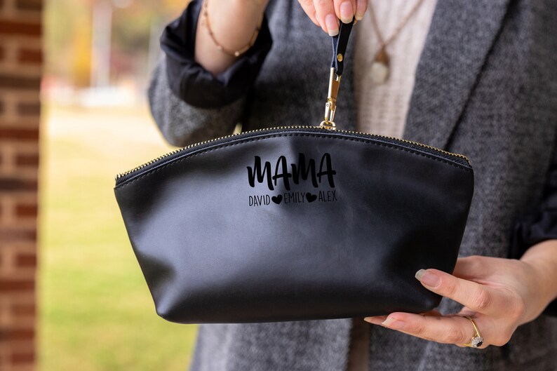 May include: Black zippered makeup bag with a gold zipper. The bag has the word "MAMA" written in a script font with three hearts and the names "DAVID", "EMILY", and "ALEX" underneath.