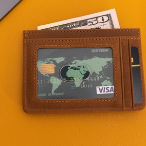 May include: A brown leather wallet with a credit card inside. The card has a world map and the Visa logo. The wallet has a slot for cash and a separate slot for cards.
