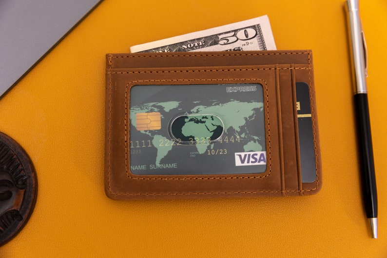May include: A brown leather wallet with a credit card inside. The card has a world map and the Visa logo. The wallet has a slot for cash and a separate slot for cards.