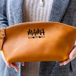 May include: A brown leather zippered pouch with the text "MAMA DAVID EMILY ALEX" in black lettering. The pouch is held in a person's hands.