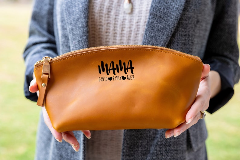 May include: A brown leather zippered pouch with the text "MAMA DAVID EMILY ALEX" in black lettering. The pouch is held in a person's hands.