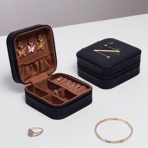 Puede incluir: Caja de joyer&iacute;a negra con cierre de cremallera y una letra dorada "N" en la parte superior. La caja est&aacute; abierta para revelar un interior marr&oacute;n con compartimentos para anillos, pendientes y otras joyas.