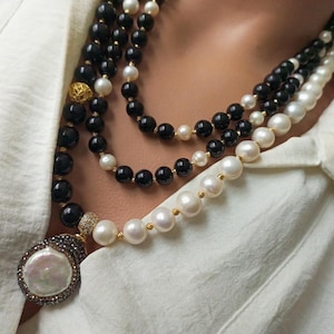 Può includere: Una collana a più fili con perline nere e bianche e un grande ciondolo. La collana presenta una combinazione di perline nere e bianche, con un grande ciondolo ornato in basso. Il ciondolo è circondato da piccole pietre scure.