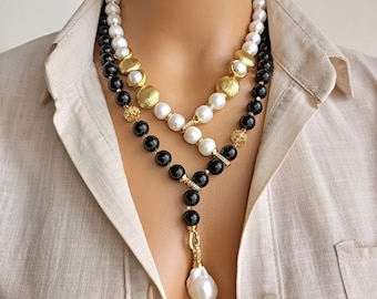 Puede incluir: Un collar de varias capas con cuentas blancas y negras, detalles dorados y un gran colgante de perla en forma de pera. El collar se muestra sobre un fondo neutro.