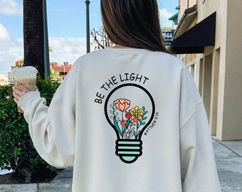 May include: A light beige sweatshirt with a light blue and white lightbulb graphic on the back. The lightbulb has flowers inside and the text "BE THE LIGHT" and "MATTHEW 5:14" on it.