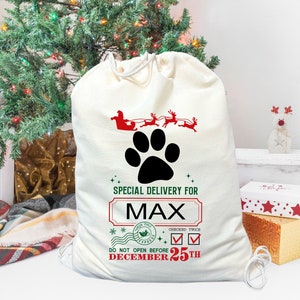 May include: A white canvas Santa sack with a black paw print, red and green text, and the name "MAX". The sack has a red and green Santa and reindeer design at the top. The text reads "Special Delivery for MAX. Checked Twice. Do Not Open Before December 25th."