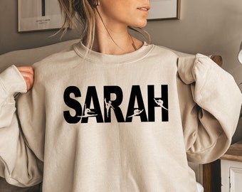 May include: A beige sweatshirt with the name "SARAH" printed in black letters. Each letter is decorated with a silhouette of a dancer in a different pose.