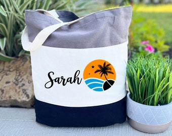 May include: A tri-colour tote bag with a grey top, white centre, and navy blue bottom. The white section features the name "Sarah" in script and a beach scene graphic with a palm tree, sun, and waves. The bag has white handles.