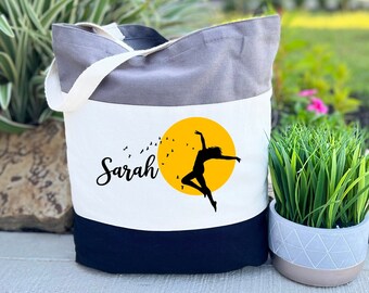 May include: A tri-color tote bag with a grey, white, and black design. The bag features a yellow sun with a silhouette of a dancer and the name "Sarah" printed on the front.